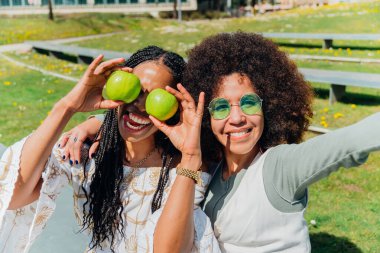 İki neşeli genç kadın parkta eğleniyor, ellerinde yeşil elmalarla dışarıda güneşli bir günün tadını çıkarırken gözlerinin önünde selfie çekiyorlar.