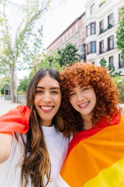 Igbtq + topluluğuna olan gurur ve desteklerini sembolize eden, canlı bir gökkuşağı bayrağına sarılmış, kucaklayan ve gülümseyen iki genç kadın, çeşitlilik ve kapsama kutlamaları sırasında