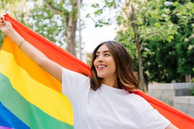Mutlu genç bir kadın beyaz bir tişört giyiyor, bir lgbtq + gurur kutlaması sırasında canlı bir gökkuşağı bayrağı sallıyor, neşeyle gülümsüyor ve uzaklığa bakıyor.