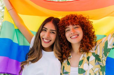 İki mutlu kadın, lgbtq + gurur, çeşitlilik ve dahil edilmeyi sembolize eden, eşit haklar için neşe ve desteği ifade eden büyük bir gökkuşağı bayrağı taşıyor.