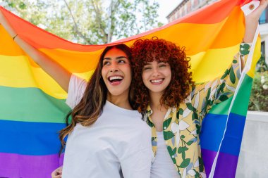 Büyük bir gökkuşağı bayrağı taşıyan iki neşeli kadın aşk ve toplum ruhuyla dolu canlı bir açık hava ortamında ışık saçan lgbt gururunu ve eşitliğini kutluyor.