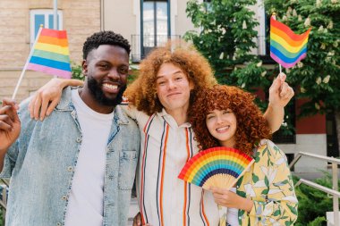 Üç mutlu çoklu etnik arkadaş gay gururunu kucaklıyor ve kutluyor, ellerinde gökkuşağı bayrakları ve bir gökkuşağı hayranıyla, lgbtq + haklarına destek veriyorlar.