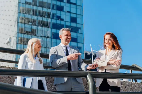 Ofis binasının önünde minyatür bir rüzgar türbini tutarken yenilenebilir enerji yatırımını tartışan takım elbiseli işadamları, kurumsal sürdürülebilirliği vurguluyorlar.
