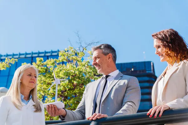 İş adamı, meslektaşlarıyla yenilenebilir enerji stratejilerini tartışırken elinde küçük bir rüzgar türbini tutuyor ve sürdürülebilir uygulamalara olan bağlılıklarını gösteriyor.