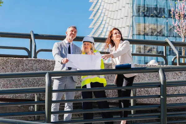 Mimarlar ve mühendislerden oluşan bir ekip, güneşli bir günde modern cam bir binanın önünde duran yeni bir inşaat projesinin planlarını inceliyor.