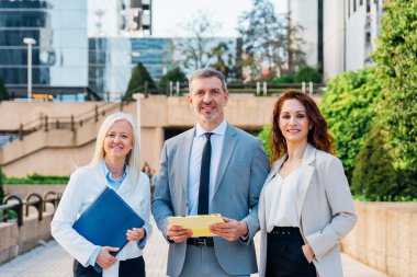 Ofis binasının önünde elinde belgeler ve dijital tabletlerle duran üç gülen iş adamı grubu başarılı işbirliklerinden memnunlar.