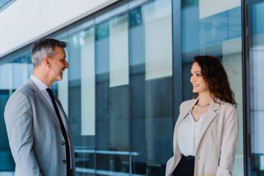 Modern bir ofis binasının önünde gülümseyen ve sohbet eden iki iş adamı canlı bir şehir ortamının ortasında arkadaşça bir iş molası veriyor.