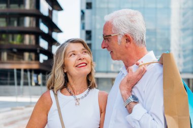 Alışveriş torbaları tutan ve birbirlerine gülümseyen neşeli yaşlı çift şehir merkezinde alışveriş çılgınlığının keyfini çıkarırken, aktif emekliliği ve tüketiciliği temsil ediyorlar.