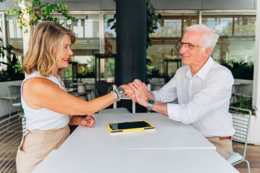 İki kıdemli iş adamı, açık hava kafeteryasında bir anlaşmayı başarıyla sonuçlandırdıktan sonra el sıkışıyor, masada bir tablet dinleniyor ve başarılarını ve ortaklıklarını vurguluyor.