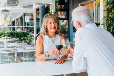 Mutlu son sınıf çifti, açık hava restoranındaki bir masada oturmuş güneşli bir günde neşe ve şefkat dolu romantik bir anın tadını çıkarırken kadehlerini şarap kadehleriyle kaldırıyor.
