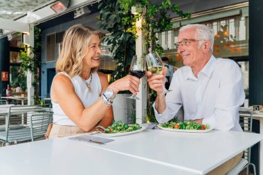 Mutlu emekli çift, açık hava restoranının verandasında sağlıklı yemeğin tadını çıkarırken kadeh kaldırıyor. Yıldönümünü ya da özel bir olayı romantik öğle yemeği ya da akşam yemeği ile kutluyor.