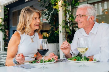 Taze salata yiyip şarap içen, açık hava restoranında sağlıklı bir öğle yemeğinin tadını çıkaran, keyifli bir sohbeti paylaşan olgun bir çift.