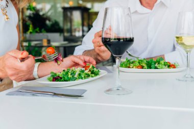 Taze salata ve şarabın yer aldığı zarif bir yemek düzeni. Sağlıklı bir yemeğin tadını çıkaran iki insan, sofistike bir ortamda malzemelerin canlılığını ve tazeliğini ön plana çıkarıyor.