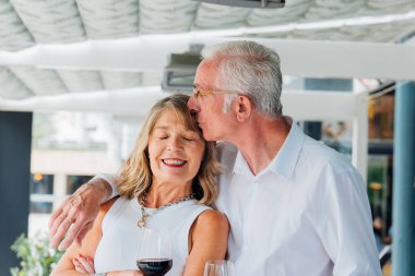 Yaşlı çift şaraptan zevk alıyor, güneşli restoran avlusunda hassas bir anı paylaşıyor, derin bir bağ ve uzun süreli romantik ilişkiyi yansıtıyor.