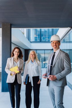 Profesyonel mühendis rüzgar türbini modelini tutuyor, modern kurumsal çalışma alanındaki iş kadınlarıyla projeyi tartışıyor, koruyucu başlık takıyor.