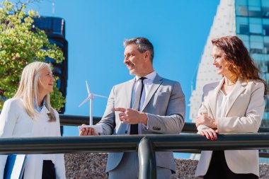 Yenilenebilir enerji yatırımını tartışan iş adamları modern ofis binalarının önünde küçük bir rüzgar türbini modeli tutuyorlar.