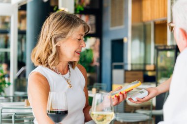 Gülümseyen olgun bir kadın, restoranın terasında elinde posizyon terminali olan bir garsonla bir bardak şarabın tadını çıkarırken temas kurmadan akıllı telefon teknolojisiyle ödeme yapıyor.