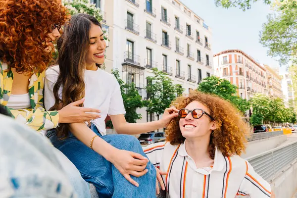 Üç mutlu genç arkadaş, iki kadın ve bir adam, alçak bir duvarda oturuyor, şehirde güneşli bir günün tadını çıkarırken gülüyor ve neşeli sohbetler ediyor.