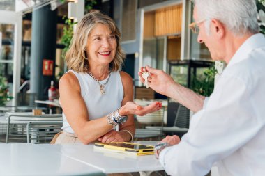 Üst düzey erkek emlakçı, açık hava bir kafede oturmuş başarılı bir emlak alımını sonuçlandırırken neşeli bir kadın müşterinin yeni evinin anahtarlarını teslim ediyor.
