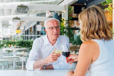 Üst düzey bir çift, açık hava restoranında şarap kadehleriyle kadeh kaldırıyor. Aşklarını ve ilişkilerini kutlarken aynı zamanda keyifli içkilerin tadını çıkarıyor ve rahatlatıcı bir atmosferde sohbet ediyorlar.