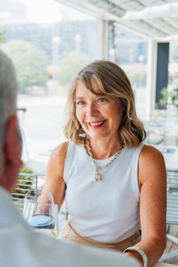 Olgun çift, samimi öğle yemeği anlarını paylaşıyor, beyaz saçlı kadın, sıcak bir şekilde gülümsüyor, dışarıda beyaz şarapla yemek yiyor.