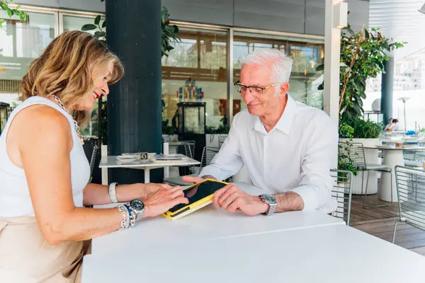 İki iş arkadaşı açık hava kafede bir tablet üzerinde iş birliği yapıyor, güneşli bir yaz gününün canlı atmosferinin tadını çıkarırken verimli bir tartışmaya giriyorlar.