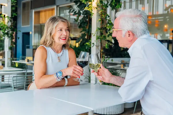 Olgun partnerler şarap kadehlerini tokuşturur, açık hava lokantasında samimi yemek deneyimlerini paylaşır, uzun süreli ilişkiyi kutlarken sıcak bir gülümsemeyle gülümserler.