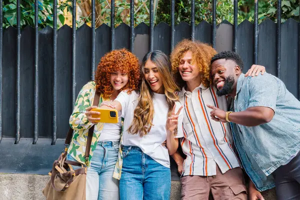 Kucaklaşan ve gülümseyen dört neşeli genç arkadaş, şehir ortamında akıllı bir telefonla selfie çekiyorlar, beraberlik ve neşe anlarını yakalıyorlar.