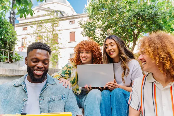Bir grup mutlu ve çeşitli üniversite öğrencisi açık havada bir proje üzerinde çalışıyorlar, dizüstü bilgisayar kullanıyorlar ve gülümsüyorlar, üniversite binasının önünde basamaklarda oturuyorlar.