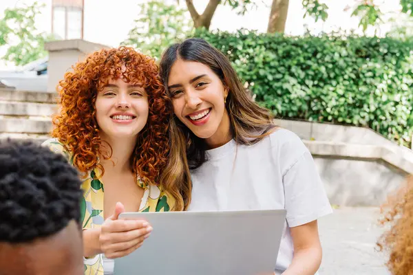 Kampüste dizüstü bilgisayarla iş birliği yaparken gülümseyen iki neşeli üniversite öğrencisi eğitim ortamlarının canlı atmosferinin ve güneşin tadını çıkarıyorlar.