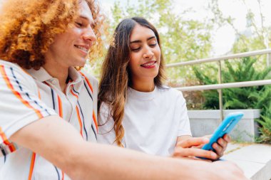 İki genç öğrenci dışarıda oturuyor, yaz havasının tadını çıkarıyor ve birlikte akıllı bir telefona bakıp gülümsüyor ve bir anlık bir bağ paylaşıyorlar.