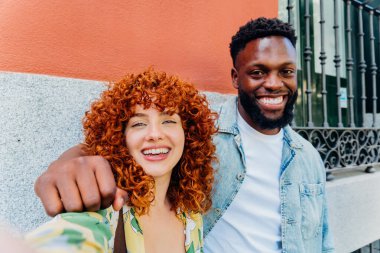 Neşeli, çoklu etnik çeşitlilikte genç bir çift, canlı bir şehir zeminine karşı selfie çekerken geniş bir gülümseme sergiliyor, neşeli bağlantılarını ve modern yaşam tarzlarını sergiliyorlar.