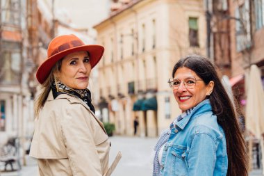 İki neşeli olgun kadın, gündelik giysiler içinde güneşli bir şehir caddesinde birlikte yürürken eğleniyorlar, gülümsüyorlar ve arkadaşlığın ve maceranın neşesini kucaklıyorlar.