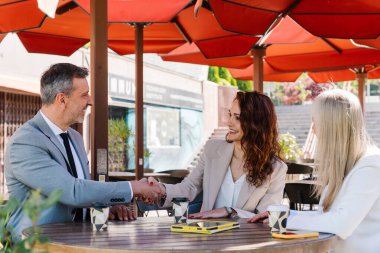 İş dünyasından profesyoneller birbirlerini selamlıyor, güneşli şehir kafesinde pazarlıkları sonuçlandırıyor, güvenilir bir el sıkışma ile başarılı bir şirket anlaşmasının sinyallerini veriyorlar.