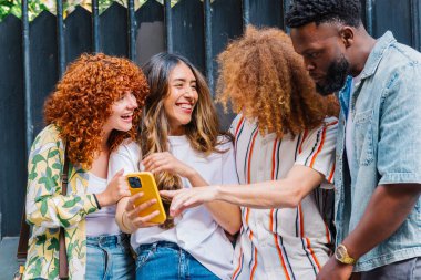 Öğrenciler ve sanatçılar da dahil olmak üzere dört neşeli genç arkadaş, dışarıda toplanmış, akıllı telefondan komik bir şeye baktıklarında bir anlık kahkaha atıyorlar.