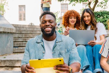 Mutlu çeşitli üniversite öğrencileri, kampüs merdivenlerinde otururken dijital tablet ve dizüstü bilgisayar kullanıyorlar, eğitim ve iletişim teknolojisinin tadını çıkarıyorlar.