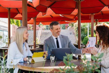 İş adamları açık havada iş birliği yapıyor, canlı kırmızı şemsiyeler altında dijital cihazlarla kafeterya masasında birlikte çalışıyor, sıcak gülümsemelerle fikir alışverişinde bulunuyorlar.