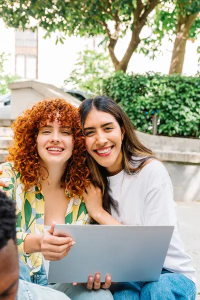 Kampüste dizüstü bilgisayar kullanırken gülümseyen ve sarılan iki genç kız öğrenci ağaçlar ve yeşilliklerle dolu canlı bir ortamda birlikte vakit geçiriyorlar.