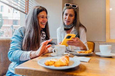 Bir kafede kahve içip kruvasan içen iki neşeli kadın sosyal medyada akıllı bir telefonla gezinirken kahkahalarını ve bağlantılarını paylaşıyor ve birlikte geçirdikleri zamanın tadını çıkarıyorlar.