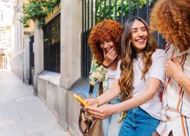 Üç neşeli genç kadın akıllı telefonlarıyla selfie çekiyor, güneşli bir günde güneşli bir caddede birlikte gülüyor ve eğleniyorlar.