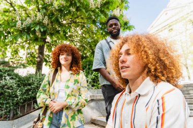 Kampüs merdivenlerinde dinlenen üç üniversite öğrencisi, canlı sohbetlere dalıyorlar ve doğa ve yeşilliklerle çevrili, canlı doğa ortamında birbirlerinin arkadaşlığından keyif alıyorlar.