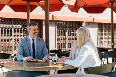 İş ortakları açık bir kafede iş projelerini tartışıyor, kahvenin tadını çıkarıyor ve kırmızı şemsiyeler altında sunumları için tablet kullanıyorlar.