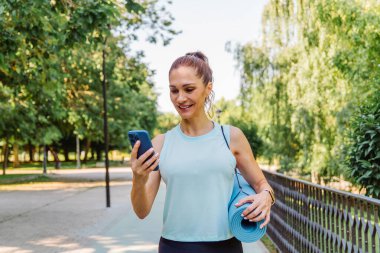 Parkta yürürken, akıllı telefonunu kullanan, yoga minderi taşıyan, sağlıklı ve aktif bir yaşam tarzının tadını çıkaran, spor kıyafeti giymiş gülümseyen bir kadın.