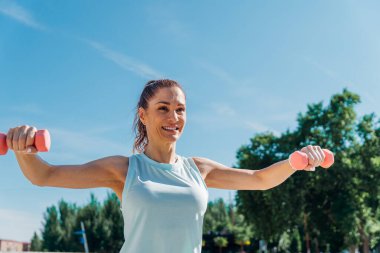 Gülümseyen sporcu kadın, açık havada pembe dambılları kaldırıyor, açık mavi gökyüzünün altında egzersiz yapıyor, yemyeşil ağaçlarla çevrili, sağlıklı ve aktif bir yaşam tarzını kucaklıyor.
