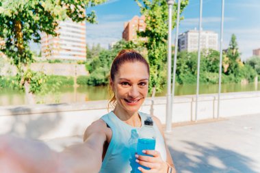 Keyifli fitness olgun bir kadın, güneşli bir yaz gününde şehirde nehir kenarında antrenmandan sonra akıllı telefonuyla selfie çekiyor, izotonik bir içecek tutuyor ve spor giysiler giyiyor.