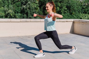 Genç sporcu kadın damdaki dambıllarla yan hamleler yapıyor, şehir manzarasının tadını çıkarırken bacaklarını ve kalçalarını güçlendiriyor.