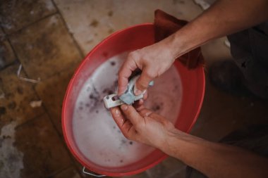 Kafkasyalı bir adamın elleri, onarımların devam ettiği bir odada çömelmiş vaziyetteki kırmızı bir kova suyun üzerinde cam çerçevesinden çıkan metal sabunla süngerle yıkanır. Üst manzara seçici bir odak noktasıyla yakın plan çekilir..