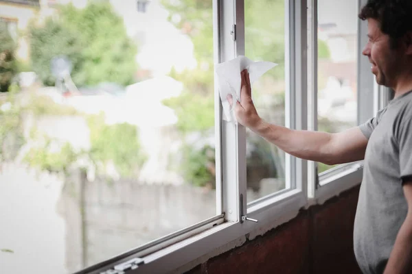 stock image Handsome caucasian man with brown curly hair with a smile on his face wipes dry paper with a napkin on a white plastic window frame, close-up side view with selective focus. The concept of repairing a house, washing window frames.