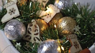 Folyo içinde yuvarlak şeker topları, yeşil süslemeli ahşap Noel oyuncakları ve beyaz bir tekstil içinde yanan bir çelenk yatak odasındaki sepetin içinde, yan görünüş yavaş çekimde..