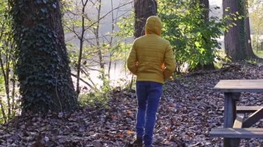 Sarı ceketli, kapüşonlu, sırtında mavi kot ve kahverengi botlarıyla genç bir adam Belçika 'nın sonbahar parkında gün batımında arka planda ağır çekimde yürür..